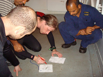 Hier laat een van onze duikers, Mitch de schade zien die hij onder water gevonden heeft. De eerste machinist, Dominic kijkt benieuwd toe. Aan de hand van de tekening kon 2 uur later een reparatie uitgevoerd worden door dezelfde duikers.
